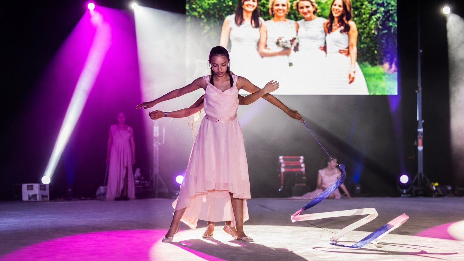 Gala de gymnastique rythmique AGTGR Avant-garde Troyes : jeune danseuse au ruban en plein numéro, écran LED en arrière-plan diffusant la photo souvenir des organisatrices en robe blanche, éclairage scénographique magenta et fumée, scène par BLK Production