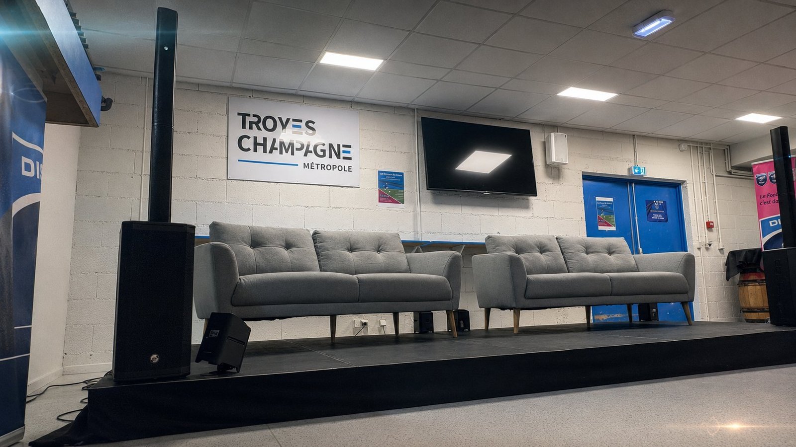 Installation scénique BLK Production dans un espace VIP de l'ESTAC Troyes : podium habillé avec deux canapés gris, sonorisation colonne haut de gamme Electro-Voice, panneau Troyes Champagne Métropole, kakemonos sportifs partenaires