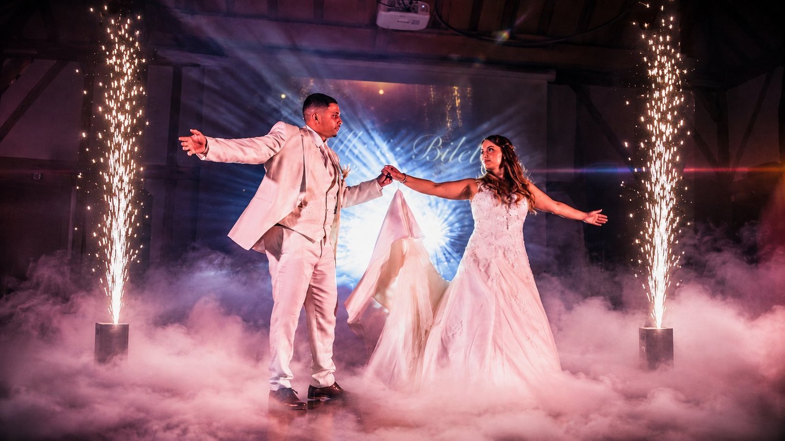 Ouverture de bal de mariage signée BLK Production, les mariés Camille et Bilel dansent au milieu d'un tapis de fumée lourde, encadrés par deux jets d'étincelles froides, avec un mapping vidéo personnalisé projeté en arrière-plan dans une grange en charpente
