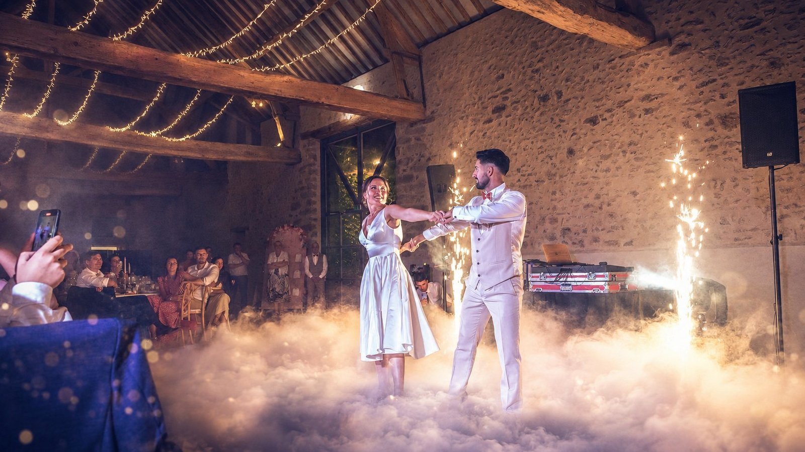 Ouverture de bal de mariage signée BLK Production dans une grange en pierre avec charpente en bois apparente, les mariés Virginie et Alexandre s'enlacent au milieu d'un tapis de fumée lourde, étincelles froides à droite, guirlandes lumineuses au plafond, invités émus qui filment la scène