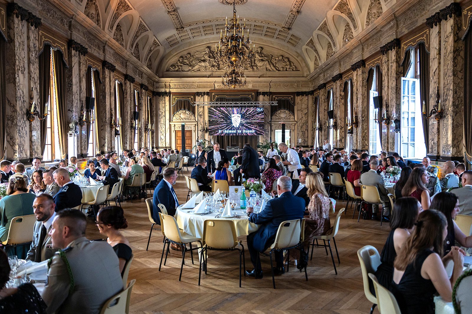 Vue d'ensemble du gala du 5e Régiment de Dragons produit par BLK Production, salle de réception historique avec lustre, colonnes de marbre et mur d'images LED aux couleurs du régiment, plusieurs centaines de convives attablés à Troyes